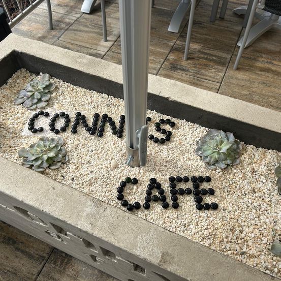Ein dekorativer Gartenbereich mit Steinen, die 'CONNES CAFE' bilden, umgeben von kleinen Sukkulenten und Kies. Dahinter befindet sich ein Holzboden mit Metallstühlen.