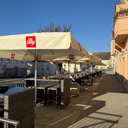 Eine Außengastronomie mit mehreren Tischen und Stühlen unter beigen Illy-Sonnenschirmen. Der Bereich befindet sich auf einem Bürgersteig mit einem Metallgeländer und Schatten auf dem Boden. Dahinter stehen Gebäude, Autos und ein Baum.