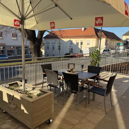 Eine Terrasse mit einem großen Sonnenschirm, einem Tisch und Stühlen. Ein Pflanzenkasten mit einem Kaffeetassendesign befindet sich links. Hinter den Gittern stehen Autos und Gebäude.