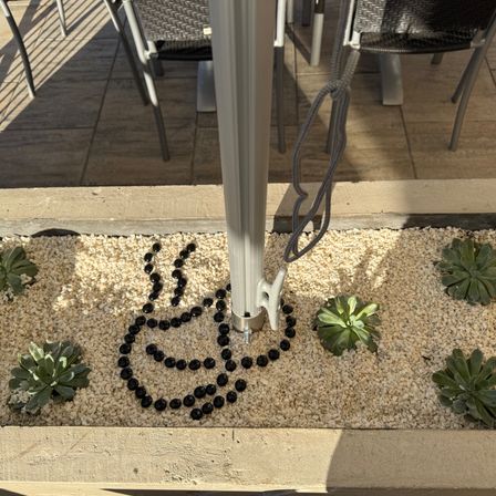 Ein dekoratives Design aus schwarzen Kieselsteinen in Herzform umgibt einen Pfosten in einem Garten. Stühle und Tische sind in der Nähe auf einem gefliesten Boden aufgestellt.