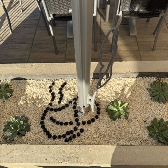 Ein dekoratives Design aus schwarzen Kieselsteinen in Herzform umgibt einen Pfosten in einem Garten. Stühle und Tische sind in der Nähe auf einem gefliesten Boden aufgestellt.