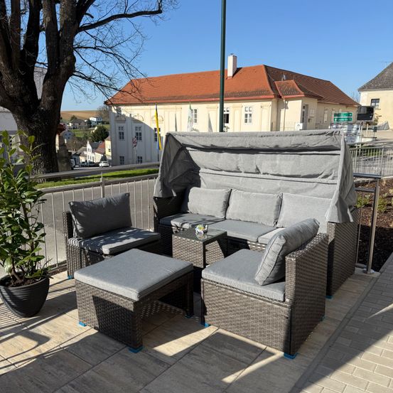 Ein Terrassenset mit überdachtem Sofa, Stühlen und einem Tisch auf einem Ziegelboden mit einem Baum und einem Gebäude im Hintergrund.