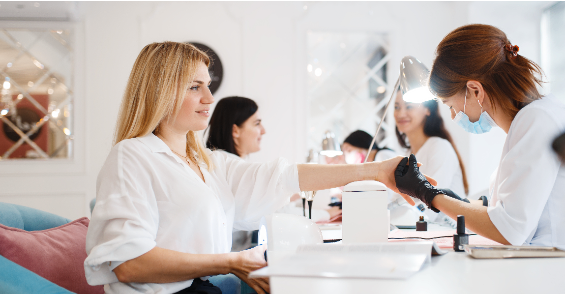 Eine Nageltechnikerin arbeitet in einem Salon und lackiert die Nägel einer Kundin, wobei eine weitere Technikerin assistiert.