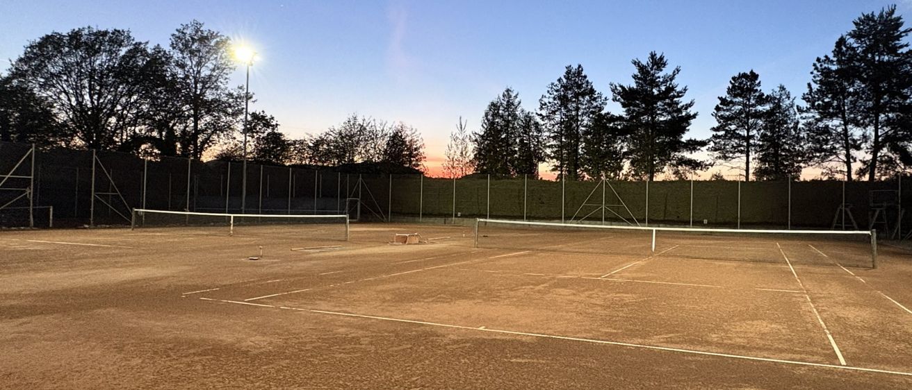 Ein leerer Tennisplatz mit zwei Netzen und zwei Straßenlaternen auf beiden Seiten, umgeben von Bäumen und einem klaren blauen Himmel.