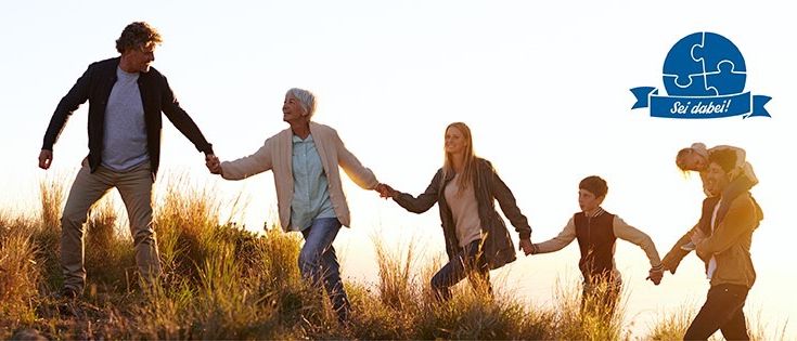 Eine Familie geht bei Sonnenuntergang in einem Grasfeld und hält Händchen. Ein älterer Mann und eine ältere Frau führen die Gruppe an. Ein Junge und ein Mädchen folgen. Ein blaues Logo mit weißem Text ist in der oberen rechten Ecke.