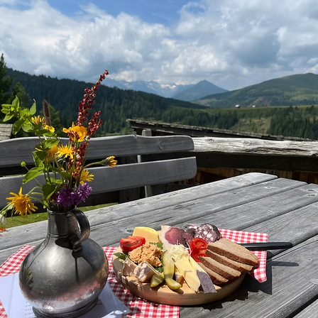 Ein Holztisch mit einer Blumenvase und einem Tablett mit Käse, Tomaten und Brot auf einem karierten Tuch, mit Blick auf eine Bergkette im Hintergrund.