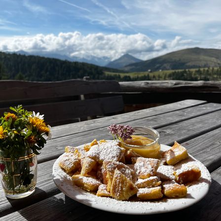 Ein Teller mit frittiertem Brot mit Puderzucker und einem kleinen Glas Marmelade auf einem Holztisch mit einer Vase voller Blumen.