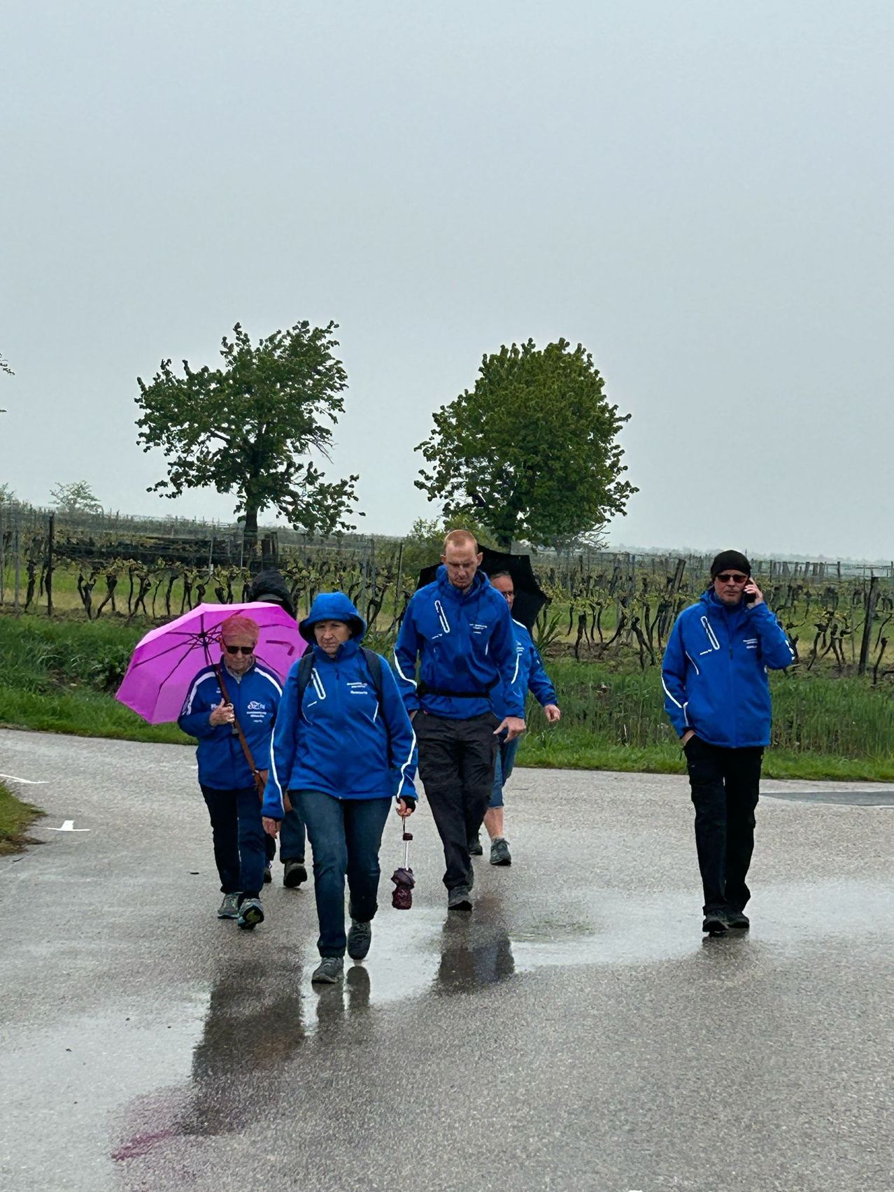 Eine Gruppe von Menschen in blauen Jacken und Hosen geht auf einer nassen Straße, zwei von ihnen halten Regenschirme und ein Mann telefoniert.