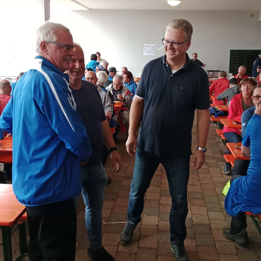 Zwei Männer stehen lächelnd in einem Speisebereich mit Ziegelfußboden. Ein dritter Mann vor ihnen führt ein Gespräch. Dahinter sitzen eine Gruppe von Menschen auf Bänken, einige tragen rote und blaue Kleidung.