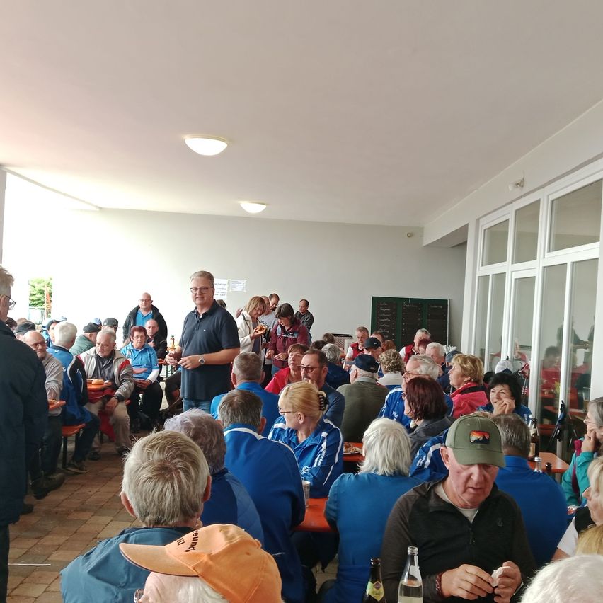 Eine Gruppe von Menschen ist in einem Speisebereich versammelt, einige stehen und andere sitzen. Sie tragen blaue Uniformen und Hüte. Ein Mann steht und scheint mit der Gruppe zu sprechen. Es gibt Flaschen und Teller auf den Tischen. Der Raum hat Glaswände und weiße Decken mit Lichtern.