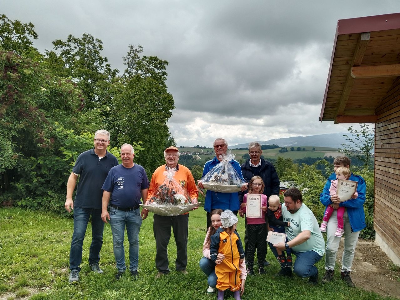 Eine Gruppe von Erwachsenen und Kindern posiert für ein Foto auf einer Grasfläche. Zwei Männer halten Blumenkörbe, während andere Urkunden halten. Im Hintergrund befinden sich Bäume und Berge.