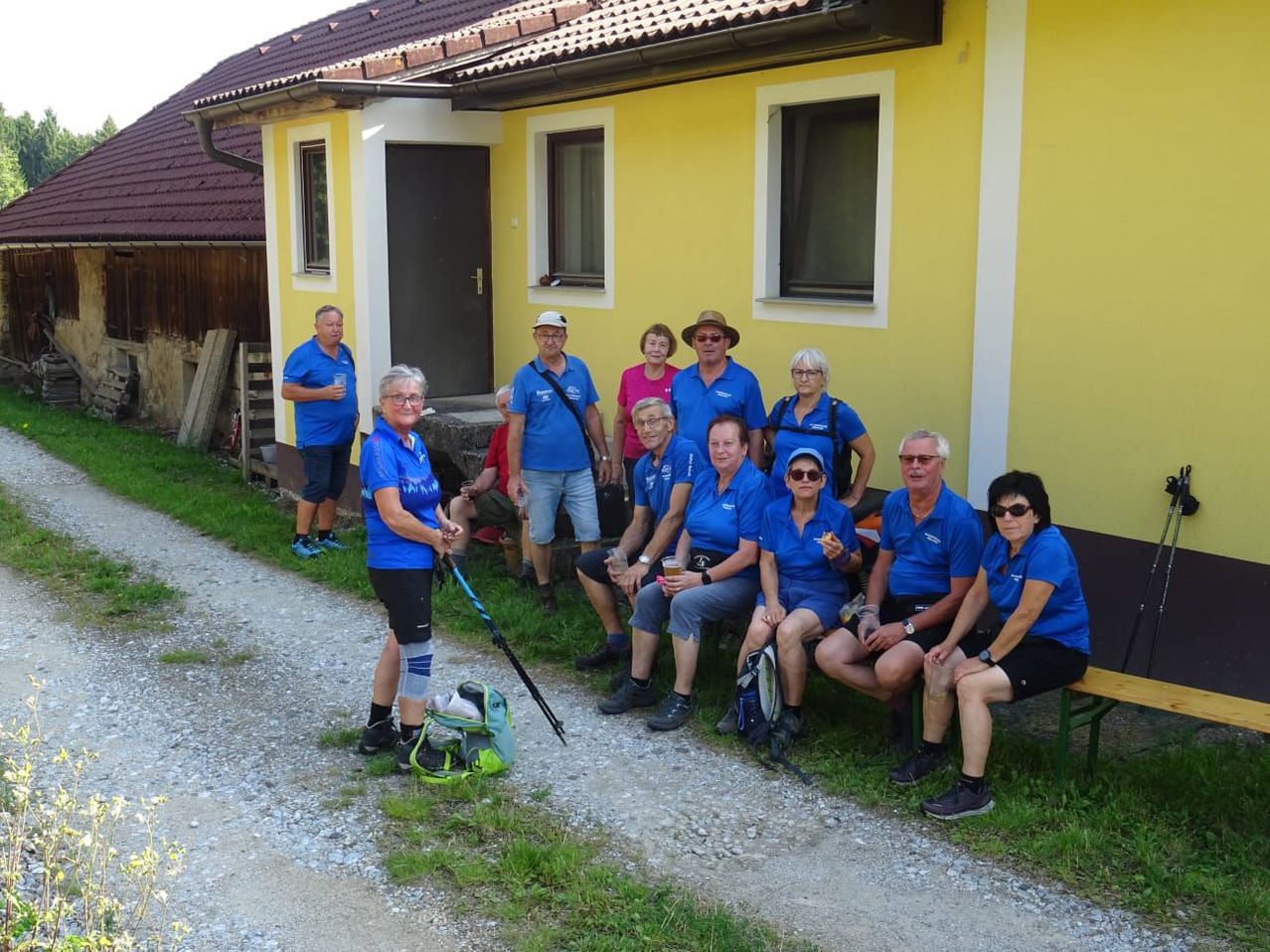 Eine Gruppe von Personen in blauen Shirts posiert für ein Foto vor einem gelben Haus. Einige sitzen auf einer Bank, während andere stehen.