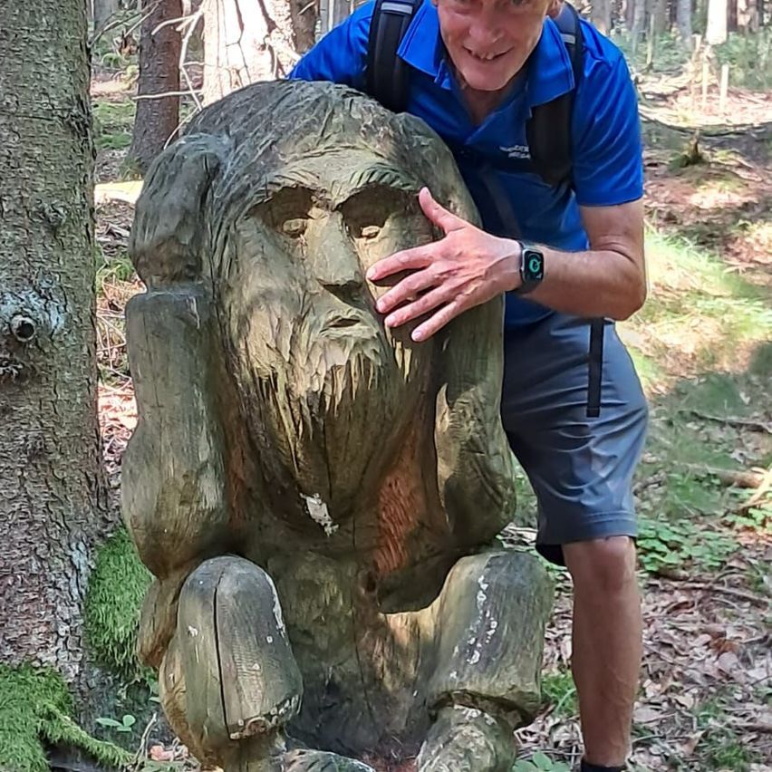 Ein Mann posiert neben einer Holzstatue eines bärtigen Mannes im Wald. Er hat einen Rucksack auf dem Rücken und trägt ein blaues Hemd.
