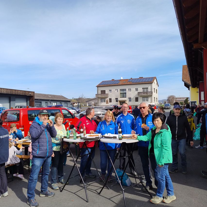 Eine Gruppe von Menschen versammelt sich um Tische auf einem Parkplatz, genießt Essen und Getränke. Sie sind in Jacken und Turnschuhen gekleidet. Im Hintergrund befinden sich Häuser, Autos und ein roter Van.