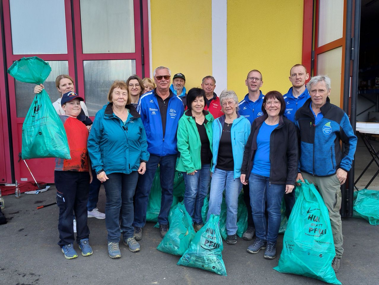 Eine Gruppe von Menschen, in verschiedenen Farben gekleidet, steht vor einem Gebäude mit gelben Wänden und roten Umrandungen. Sie halten grüne Taschen.