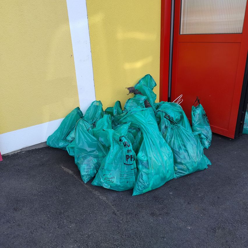 Mehrere grüne Plastiktüten sind vor einer roten Tür mit Fenster auf einem gelben Wandstapel gestapelt.