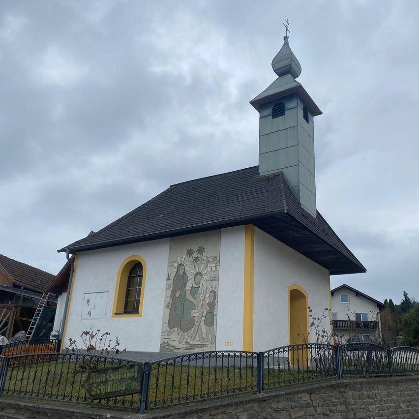 Eine kleine Kirche mit schwarzem Dach und Turm steht gegen einen bewölkten Himmel. Die Kirche hat eine große Wandmalerei auf ihrer Seite und gelbe Verzierungen um die Fenster und Türen. Ein Zaun umgibt die Kirche.