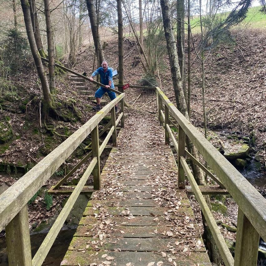 Ein Mann in einer blauen Jacke steht auf einer hölzernen Brücke mit Geländer in einem Wald. Die Brücke überquert einen Bach mit Felsen und gefallenen Blättern. Bäume umgeben das Gebiet, und der Boden ist uneben.