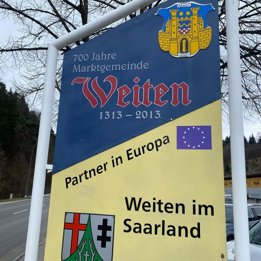 Ein blaues und gelbes Schild steht draußen und zeigt '700 Jahre Marktgemeinde Weiten 1313 - 2013.' Darunter steht 'Partner in Europa Weiten im Saarland' mit einem EU-Emblem.