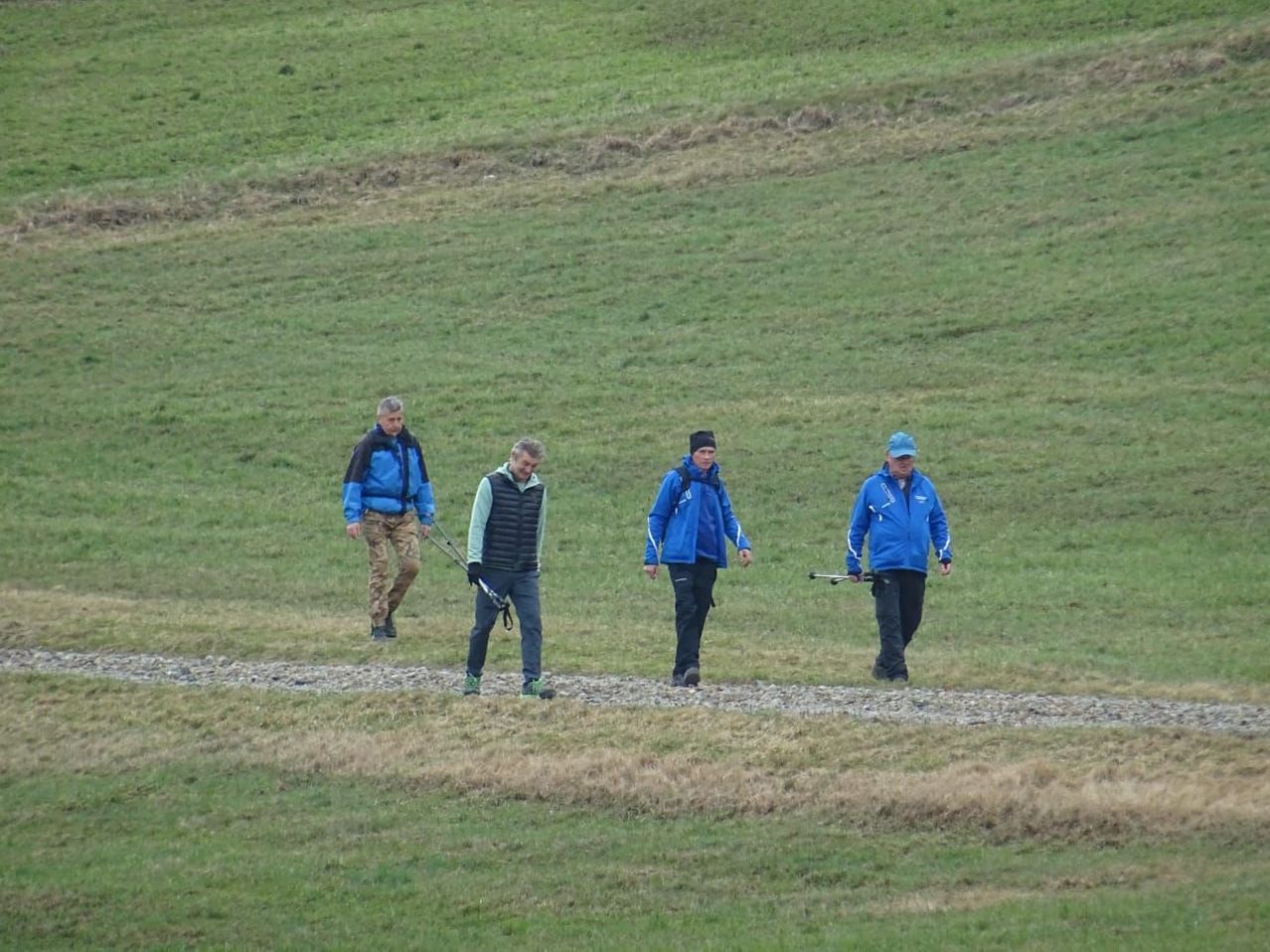 Vier Personen gehen auf einem Kiesweg durch ein Grasfeld. Sie tragen alle blaue Jacken und Hosen, und einige haben Hüte auf. Sie scheinen einer Freizeitaktivität nachzugehen.