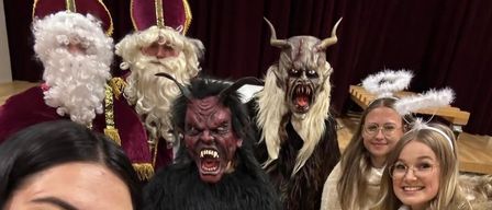 Eine Gruppe von Menschen, darunter Frauen in Kostümen, posiert für ein Foto. Eine Frau ist als Weihnachtsmann gekleidet, während eine andere als Dämon verkleidet ist. Sie stehen auf einem Holzboden.