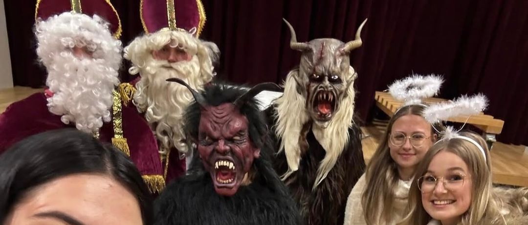 Eine Gruppe von Menschen, darunter Frauen in Kostümen, posiert für ein Foto. Eine Frau ist als Weihnachtsmann gekleidet, während eine andere als Dämon verkleidet ist. Sie stehen auf einem Holzboden.