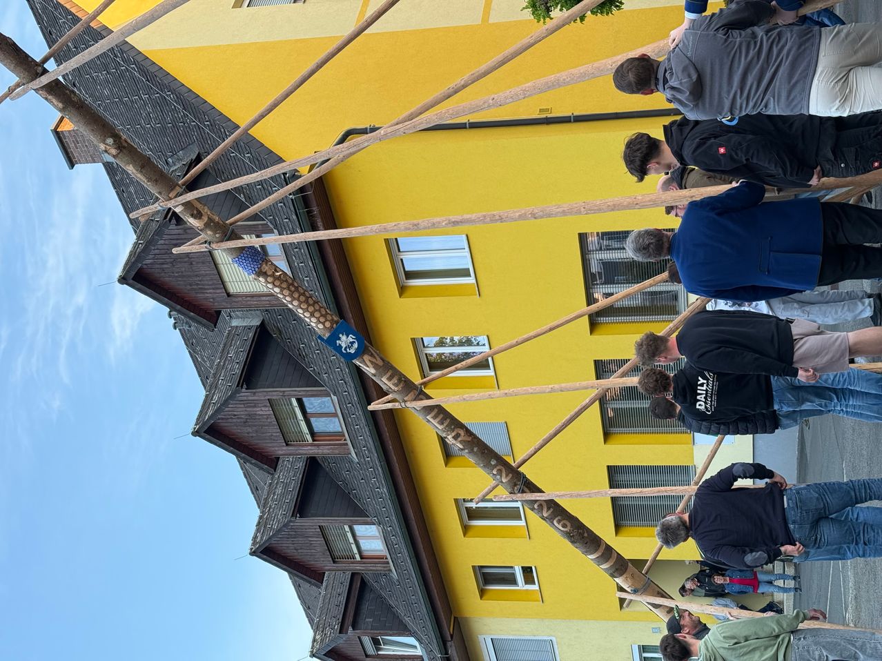 Eine Gruppe von Menschen versammelt sich um eine Holzstruktur, möglicherweise ein Maibaum, vor einem gelben Gebäude mit mehreren Fenstern.