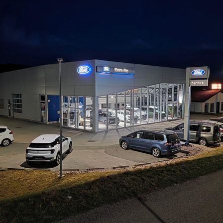 Ein Ford-Autohaus bei Nacht mit mehreren Autos vor dem Gebäude geparkt, beleuchtet durch Außenbeleuchtung. Das Gebäude hat ein modernes Design mit Glaswänden und einem blauen Ford-Schild.