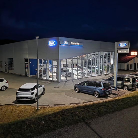 Ein Ford-Autohaus bei Nacht mit mehreren Autos vor dem Gebäude geparkt, beleuchtet durch Außenbeleuchtung. Das Gebäude hat ein modernes Design mit Glaswänden und einem blauen Ford-Schild.