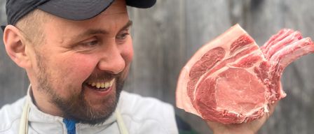 Ein Mann in einer schwarzen Kappe und einem weißen Lendenschurz lächelt und hält ein Stück rohes Fleisch.