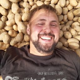 Ein lächelnder Mann liegt auf einem Bett aus Kartoffeln, trägt ein schwarzes T-Shirt mit weißem Text und ist von einer Fülle von Kartoffeln umgeben.