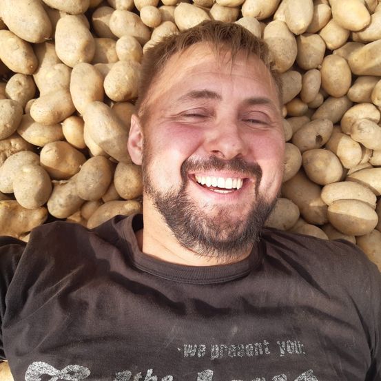 Ein lächelnder Mann liegt auf einem Bett aus Kartoffeln, trägt ein schwarzes T-Shirt mit weißem Text und ist von einer Fülle von Kartoffeln umgeben.