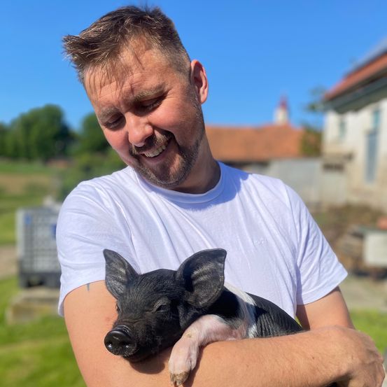 Ein Mann in einem weißen T-Shirt hält ein schwarz-weißes Ferkel und lächelt. Im Hintergrund befindet sich ein Rasen mit Gebäuden und Bäumen.