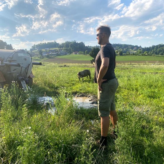 Ein Mann steht in einem Grasfeld, mit einem Schwein und einem großen Maschinenfahrzeug im Hintergrund, unter einem bewölkten Himmel.