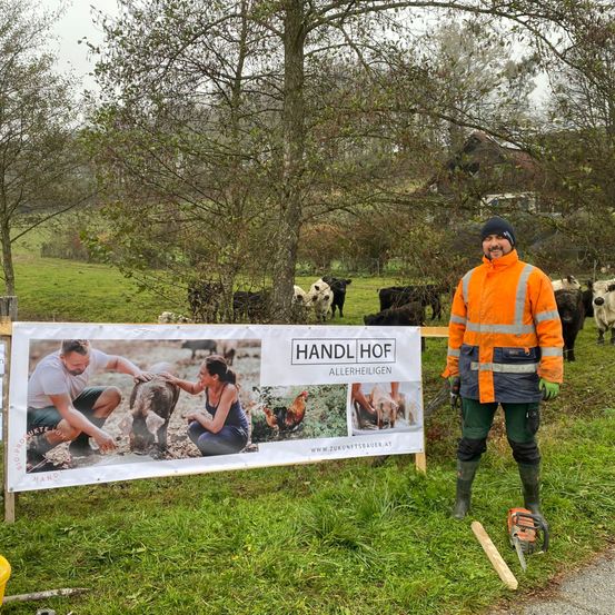 Ein Mann steht neben einem Banner, das Menschen mit Nutztieren zeigt, und posiert für ein Foto auf einem Bauernhof mit Kühen und einem Haus im Hintergrund.