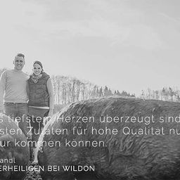 Ein Paar steht lächelnd neben einem Schwein. Im Hintergrund ist ein Wald zu sehen. Rechts hockt ein Mann und untersucht einen Stein, mit deutschem Text über Qualität aus der Natur.