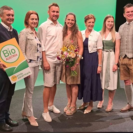 Eine Gruppe von Menschen steht auf einer Bühne und lächelt für die Kamera. Ein Mann hält ein Schild mit der Aufschrift 'Bio Austria'. Die Gruppe besteht aus Männern und Frauen mit verschiedenen Schuhen und Outfits. Eine Frau in der Mitte hält einen Blumenstrauß.