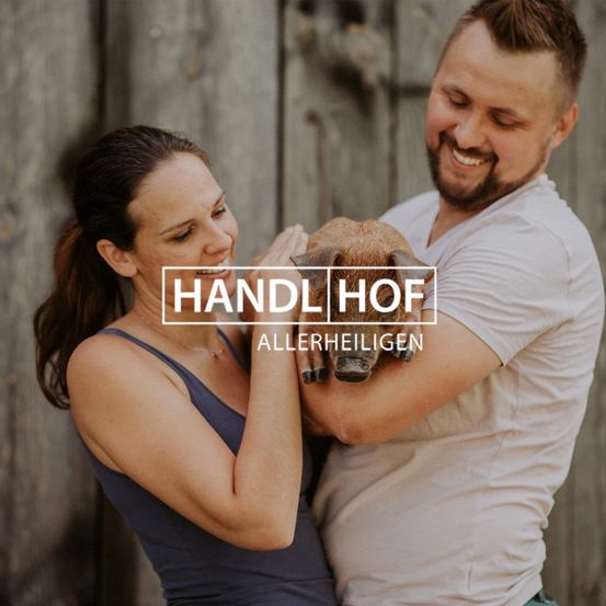 Ein lächelndes Paar steht mit einem kleinen Schwein, das sie zwischen sich halten. Sie sind in der Nähe einer Holzzaun. Der Text lautet 'HANDL HOF ALLERHEILIGEN'.