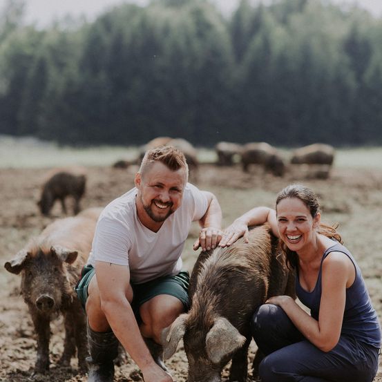Ein lächelndes Paar kniet neben einem braunen Schwein in einem schlammigen Bereich, mit anderen Schweinen und Bäumen im Hintergrund.