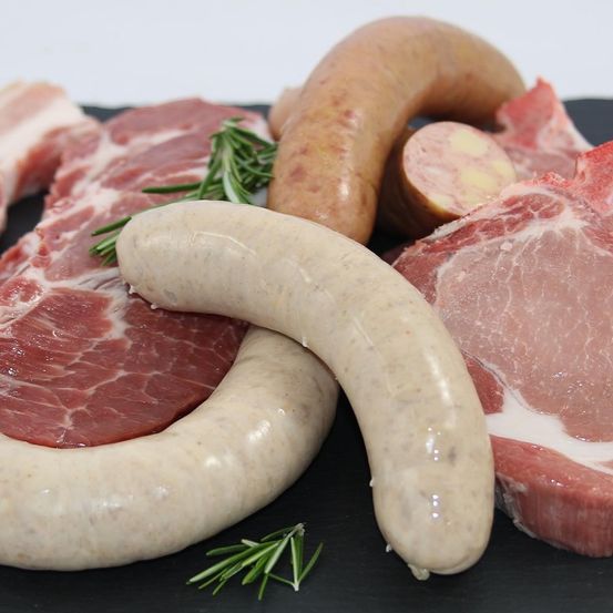 Eine Auswahl an frischem Fleisch und Würsten, einschließlich eines Fleischstücks mit Rosmarin, zwei Würsten und einem Stück Speck, liegt auf einer schwarzen Oberfläche.