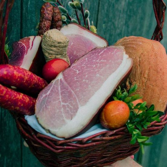 Ein Korb gefüllt mit Wurst, Schinken, Brot und Früchten, auf einem grünen Hintergrund platziert.