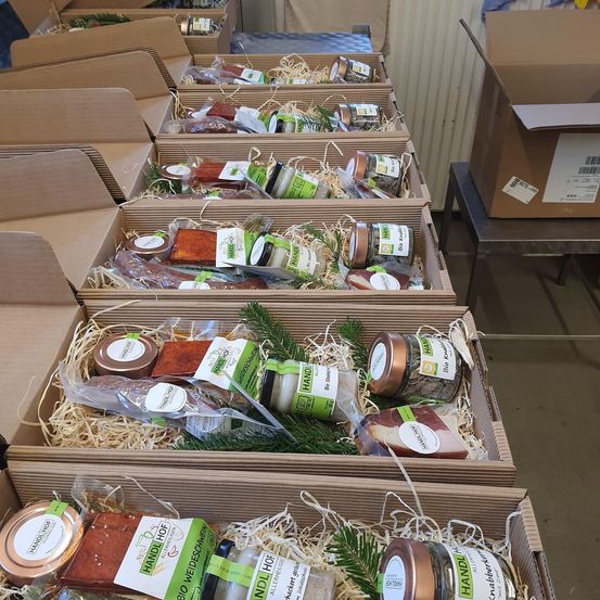Mehrere Kisten mit verpackten Lebensmitteln sind auf einem Tisch ausgestellt. Jede Kiste enthält eine Vielzahl von Produkten, einschließlich Gläser und Fleischscheiben, die in Plastik eingewickelt und mit Heu gepolstert sind. Die Etiketten auf den Kisten zeigen an, dass sie versandfertig sind.