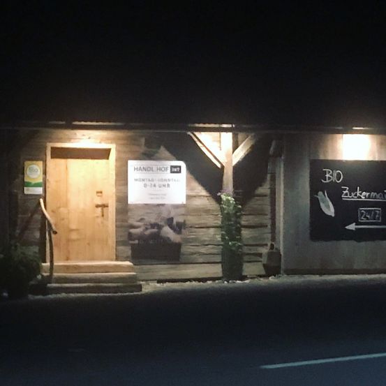 Ein hölzernes Gebäude wird bei Nacht gezeigt mit einem beleuchteten Schild, das 'BIO Zuckermais 24/7' anzeigt. Das Gebäude hat eine Holztür mit einem Griff und eine hölzerne Treppe. Ein Topf mit einer Pflanze ist auf der Seite des Gebäudes platziert.