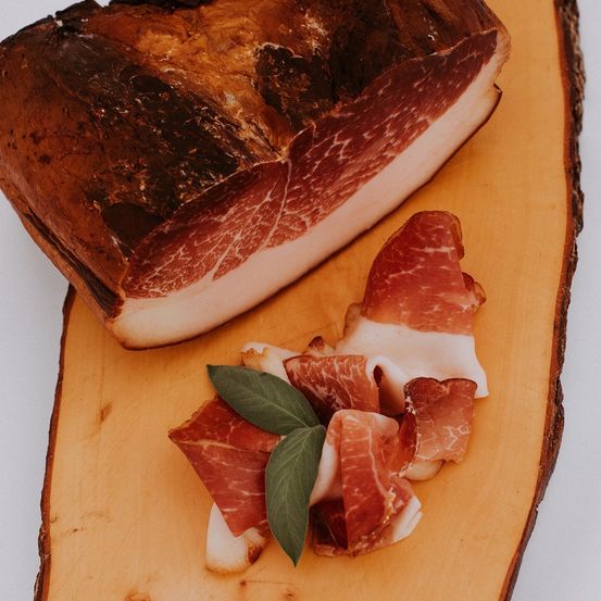 Ein großes Stück gepökeltes Fleisch liegt auf einem hölzernen Schneidebrett, begleitet von kleineren Scheiben und einem grünen Blatt.