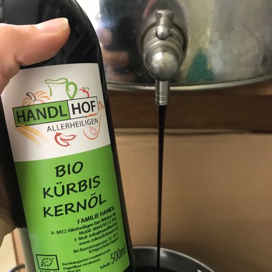 Eine Hand hält eine Flasche Bio-Kürbiskernöl und gießt es in einen Topf. Das Etikett zeigt 'BIO KURBIS KERNOEL'.
