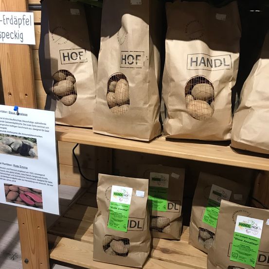Ein Holzregal hält mehrere braune Kartoffelsäcke. Jeder Sack ist mit dem Wort 'HOF' oder 'HANDL' beschriftet und hat ein kleines Fenster, das die Kartoffeln im Inneren zeigt. Ein weißes Schild auf der linken Seite des Regals zeigt Text.