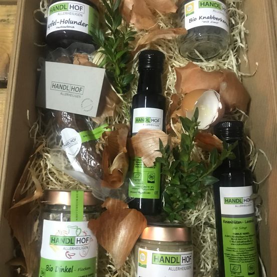 Eine Box mit verschiedenen Produkten von 'Handl Hof', darunter Gläser, Flaschen und Tüten, auf einem Heu- und Laubbett angeordnet, mit einem zerbrochenen Eierschalenstück.