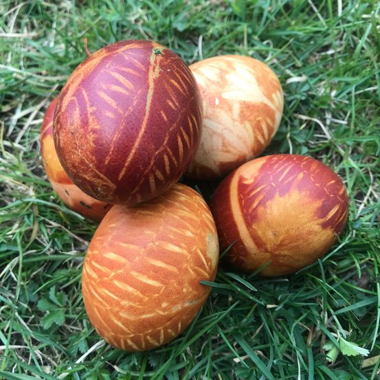Vier kunstvoll dekorierte Eier mit rotbraunen Mustern liegen auf einer Grasfläche. Die Muster ähneln Blättern und sind detailreich.