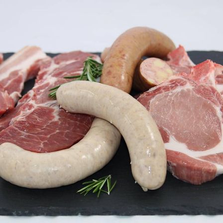 Eine Auswahl an rohem Fleisch und Würsten ist auf einem schwarzen Schieferbrett angeordnet, einschließlich Scheiben von Schweinefleisch, Rindfleisch und Würsten mit Rosmarin.