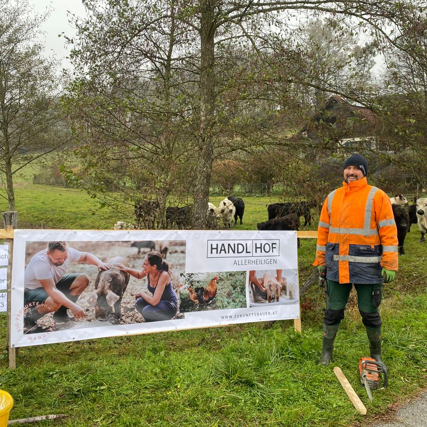 Ein Mann in einer orangefarbenen Weste steht neben einem Schild auf einem Bauernhof, im Hintergrund sind Kühe und Hühner zu sehen.